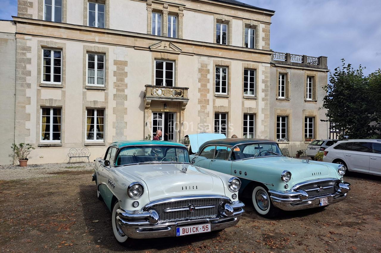 Deux voitures anciennes stationnées devant un bâtiment historique en pierre, sous un ciel légèrement nuageux.