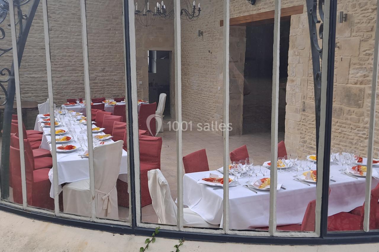 Tables dressées avec nappes blanches et chaises rouges dans une salle en pierre, vue à travers des barreaux métalliques.