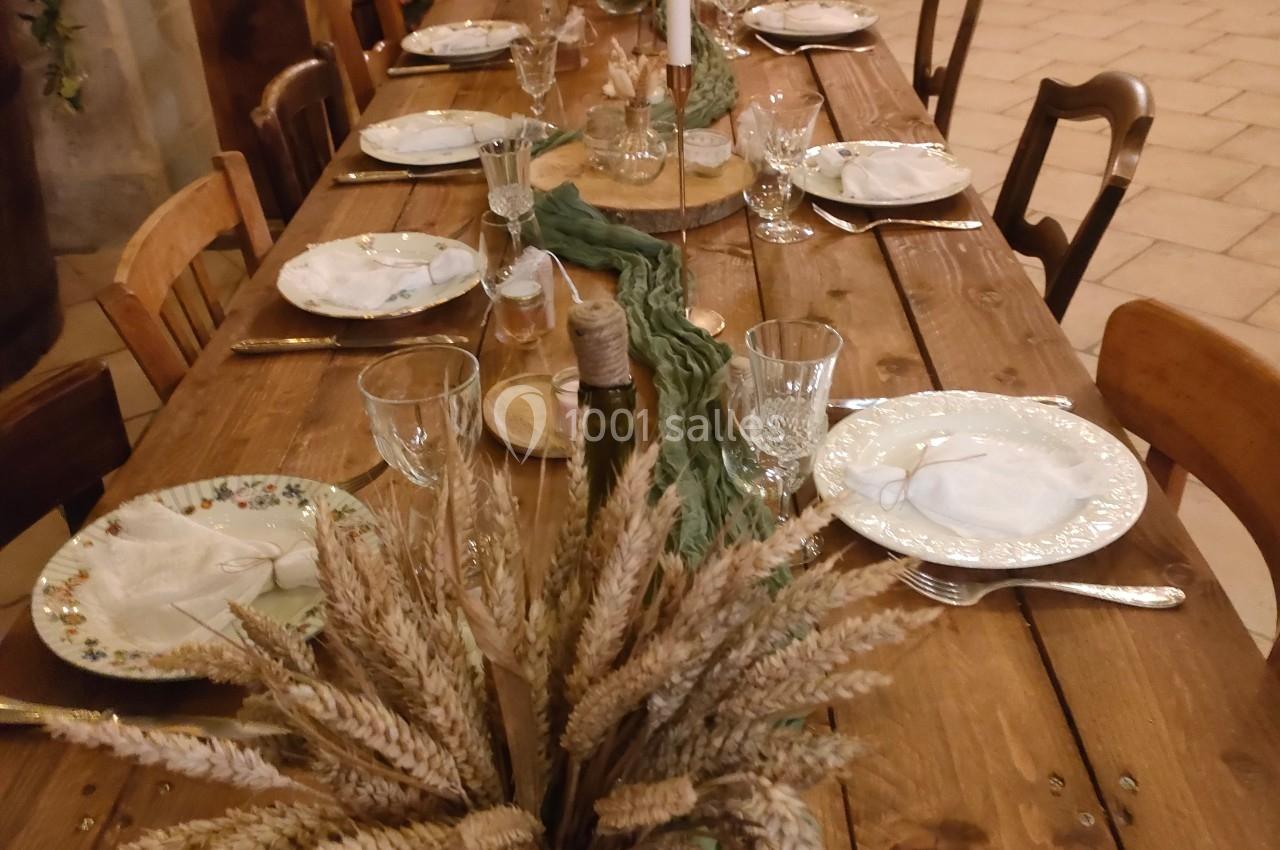 Table en bois décorée pour un dîner, avec assiettes, verres, bougies et centre de table en épis de blé et tissu vert.