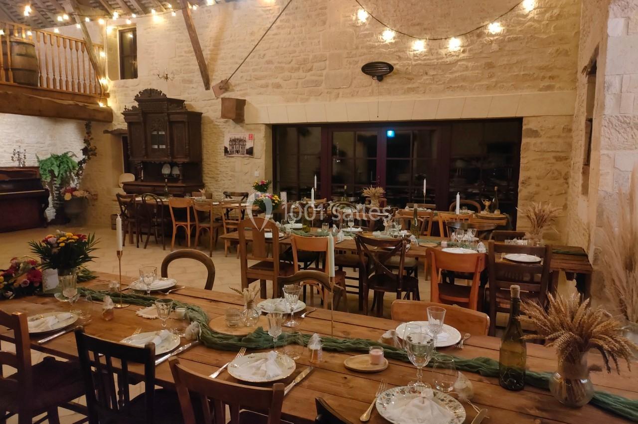 Salle de réception rustique avec tables en bois décorées, chaises variées et guirlandes lumineuses suspendues.