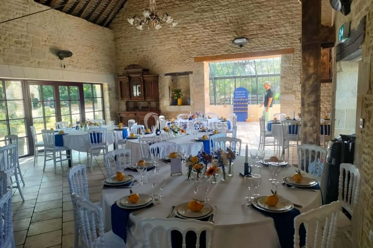 Salle de réception lumineuse avec tables dressées, décorées de fleurs et serviettes bleues, dans un cadre en pierre.