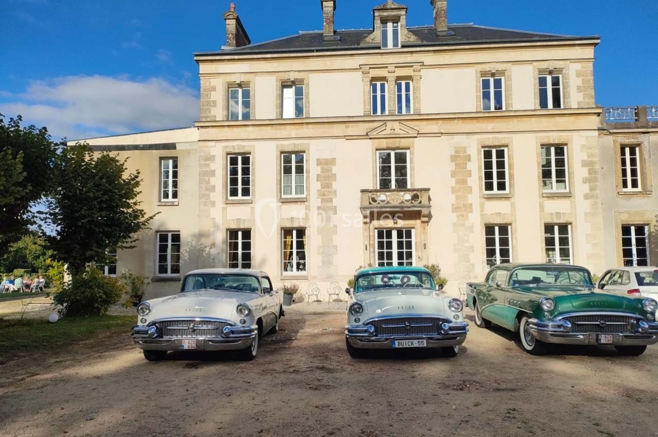 Façade d'un manoir en pierre avec quatre voitures anciennes stationnées devant sur un terrain ensoleillé.
