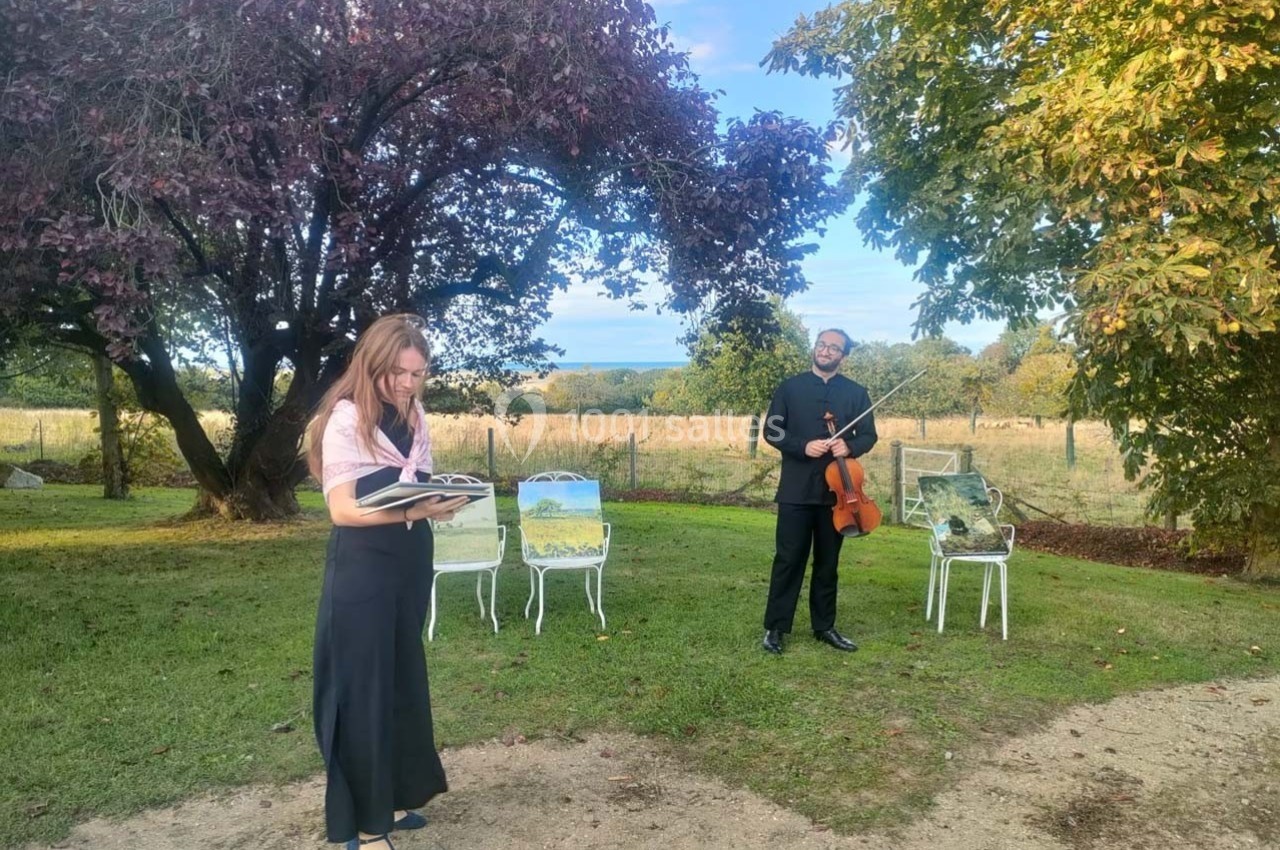 Une femme lit un livre et un homme tient un violon dans un jardin, avec des tableaux posés sur des chaises.