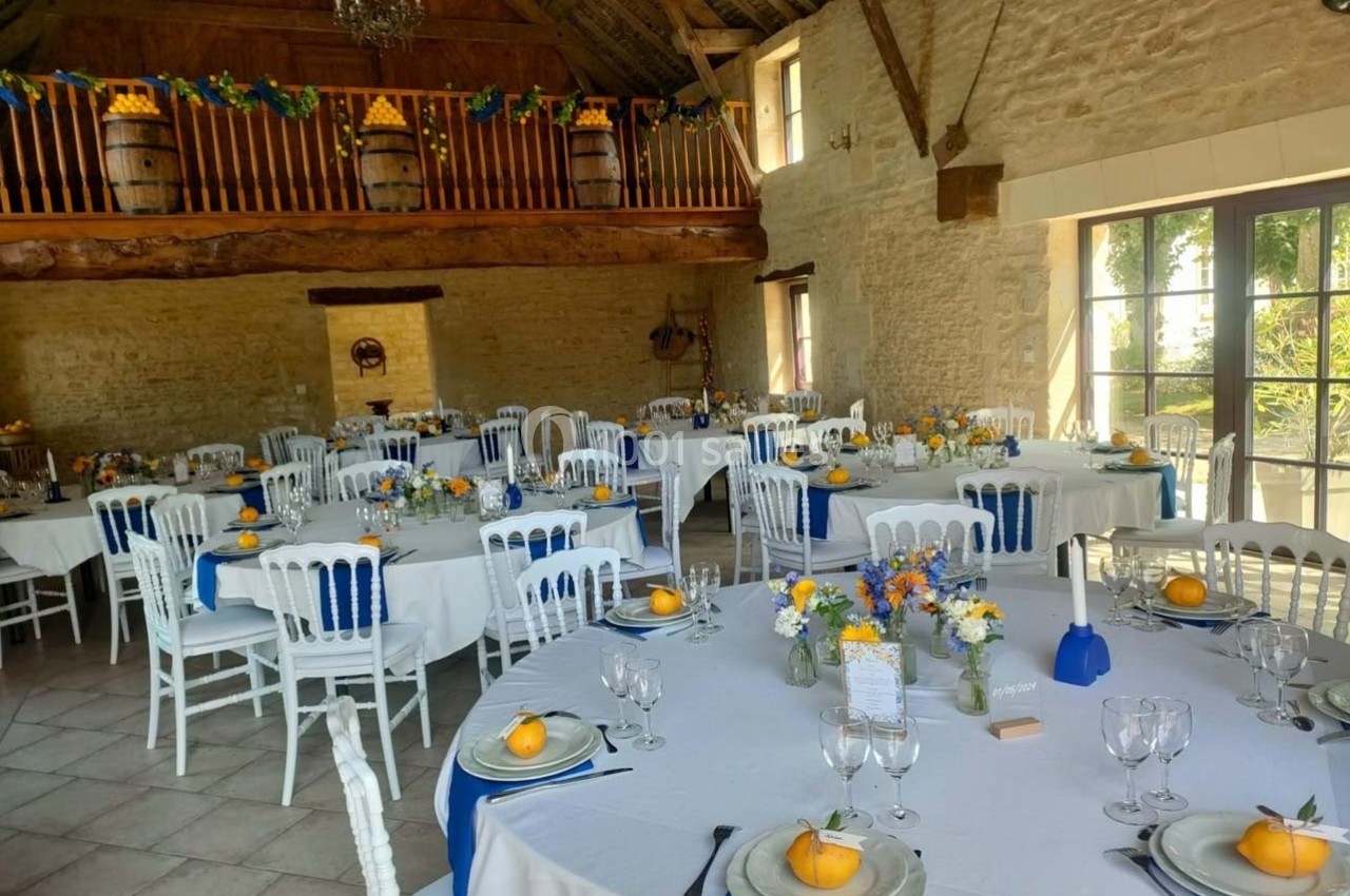 Salle de réception rustique décorée avec des tables rondes, nappes blanches, citrons en centre de table et poutres…