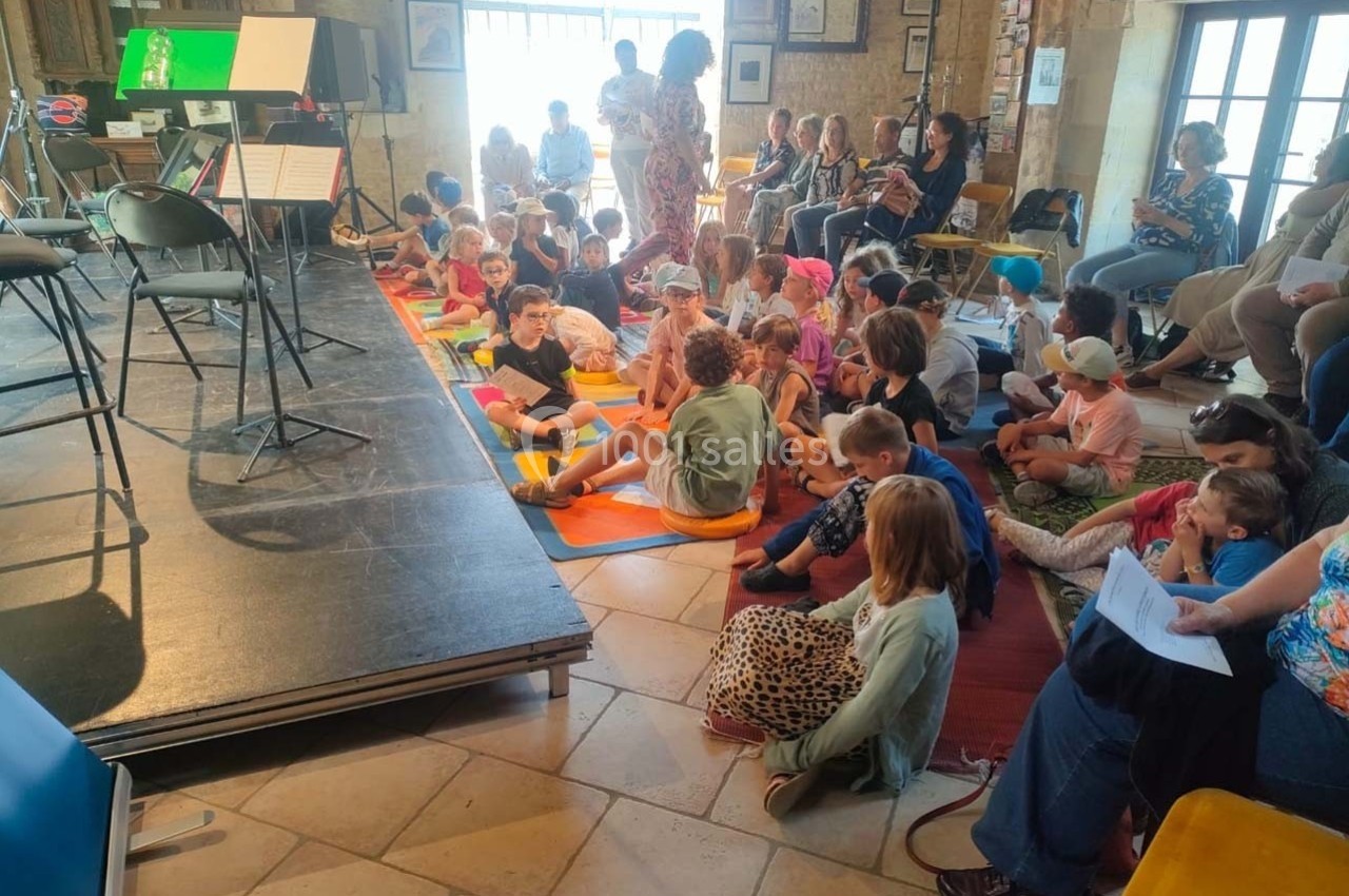 Des enfants assis sur des tapis colorés assistent à une représentation musicale dans une salle avec un public varié.