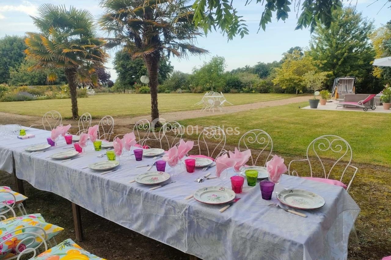 Table dressée en extérieur avec nappes blanches, assiettes, verres colorés et serviettes roses, entourée d'un jardin…