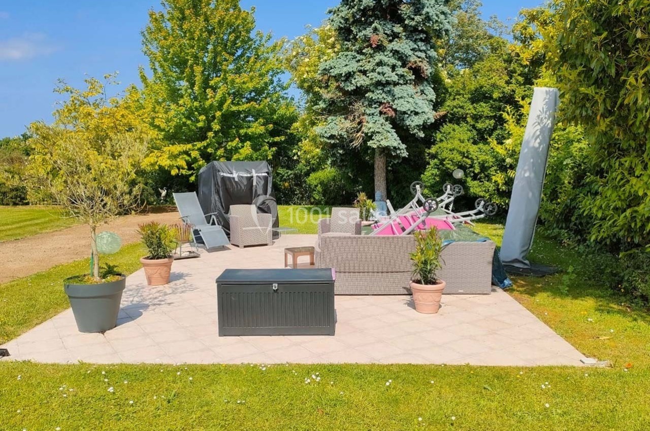 Terrasse extérieure avec mobilier de jardin en rotin, plantes en pot et arbres en arrière-plan par temps ensoleillé.