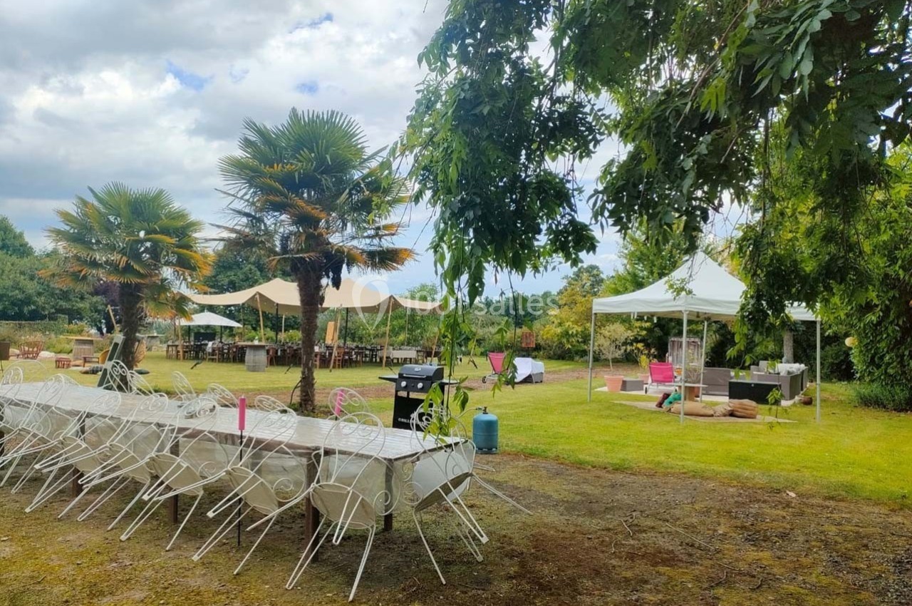 Tables et chaises blanches disposées en extérieur sous des arbres, avec des tentes et un espace aménagé dans un jardin.