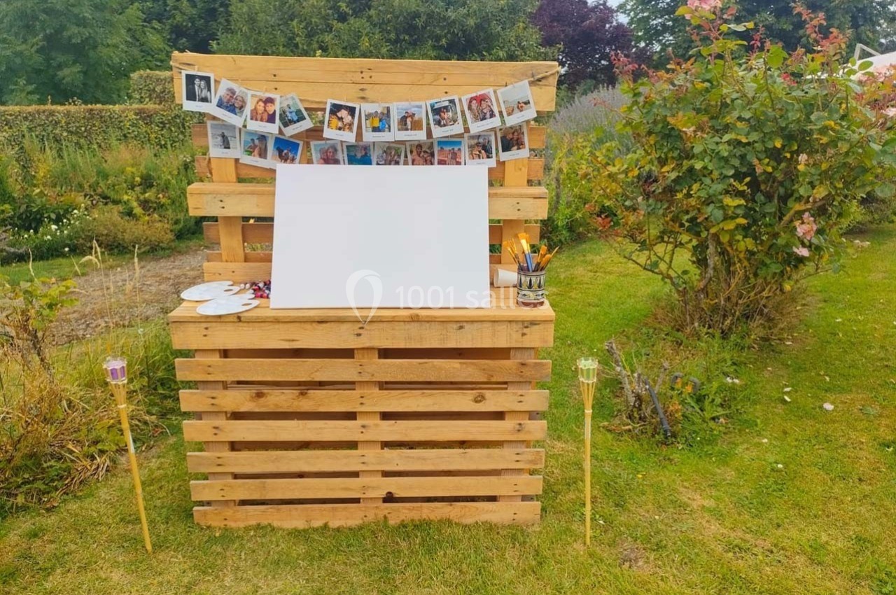 Stand en palettes avec une toile blanche, guirlande de photos, pinceaux et crayons, installé dans un jardin fleuri.