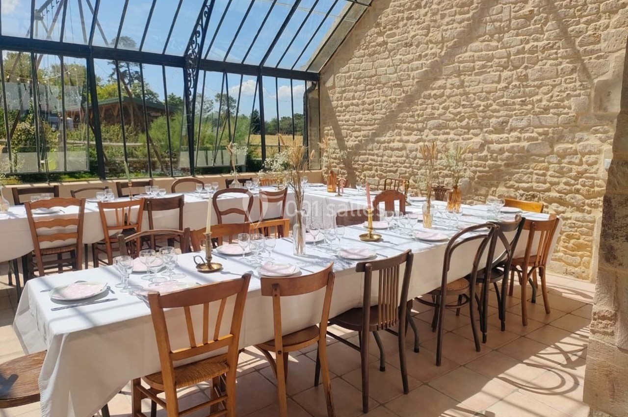 Salle lumineuse avec de grandes baies vitrées, tables dressées avec nappes blanches et chaises dépareillées.