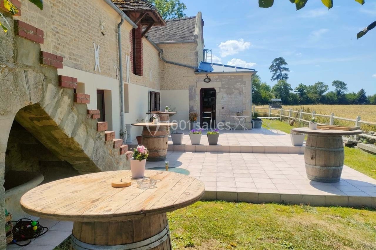 Terrasse ensoleillée avec tables en tonneaux, fleurs en pot et vue sur un champ bordé d'arbres.