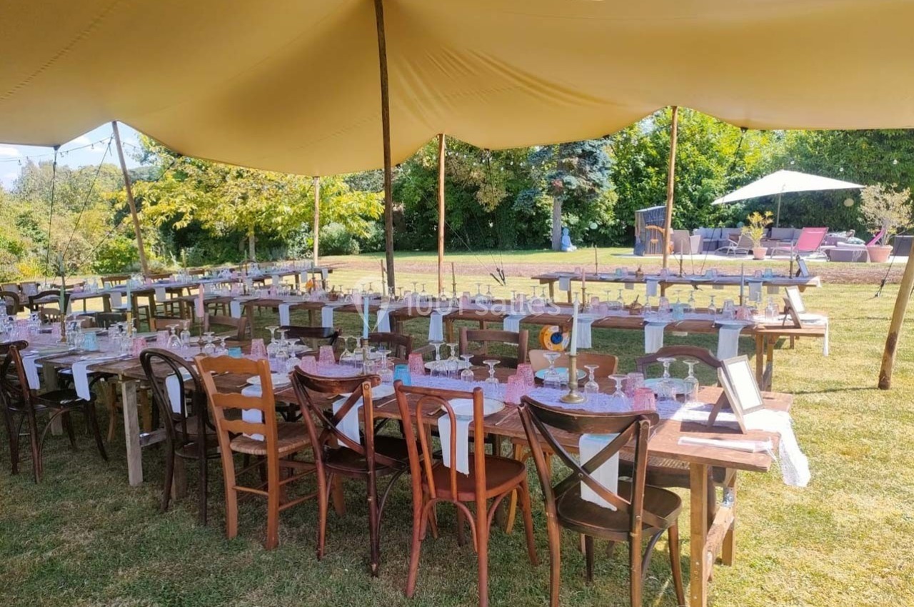 Tables en bois décorées pour un repas en extérieur sous une tente, avec chaises dépareillées et vue sur un jardin.