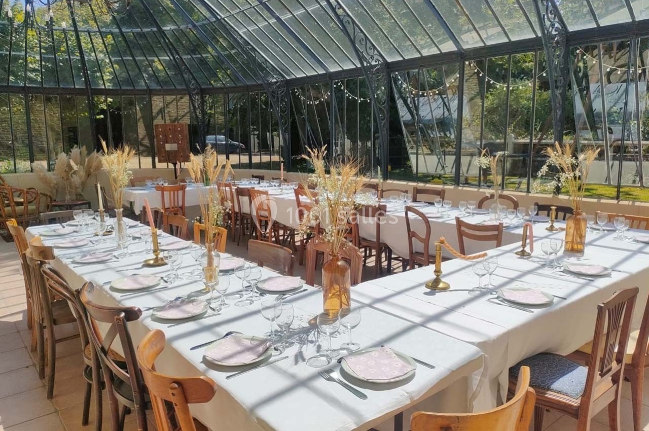 Tables dressées avec nappes blanches, vaisselle et décorations florales dans une verrière lumineuse avec vue sur un jardin.