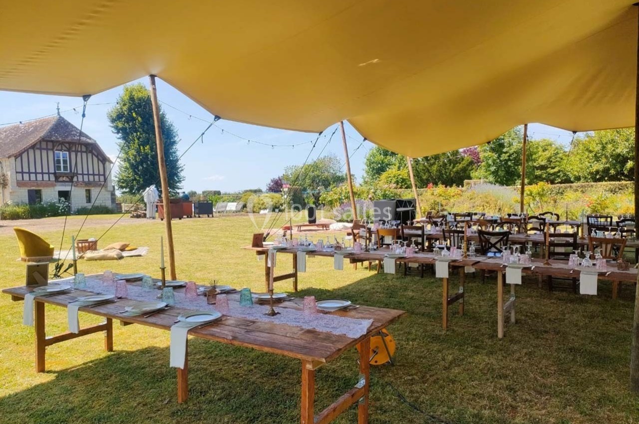Tables en bois dressées sous une tente beige dans un jardin, avec une maison à colombages en arrière-plan.