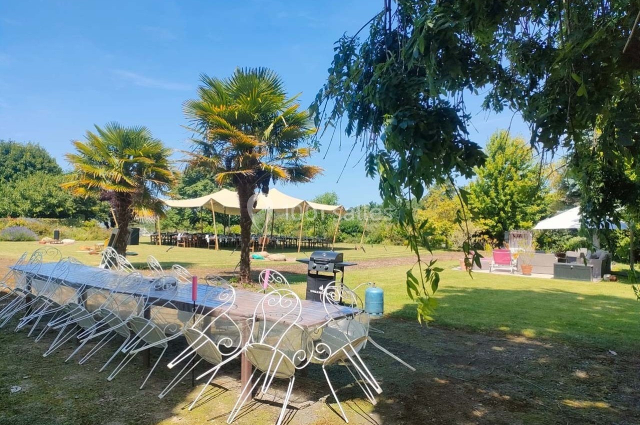 Tables et chaises blanches disposées en extérieur sous des arbres et des parasols, dans un jardin ensoleillé.