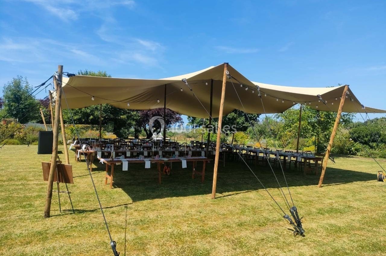 Tente beige montée sur un terrain herbeux, abritant des tables dressées pour un événement en plein air.