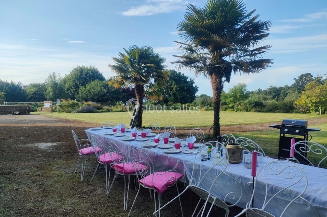 Table dressée en extérieur avec chaises blanches et coussins roses, sous des palmiers, dans un jardin verdoyant.