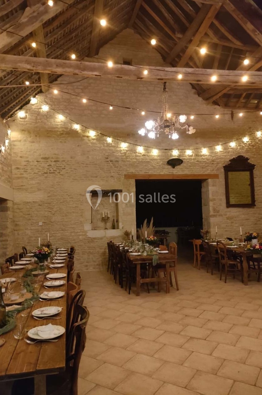 Salle en pierre avec tables dressées, guirlandes lumineuses suspendues et ambiance chaleureuse pour un repas ou événement.