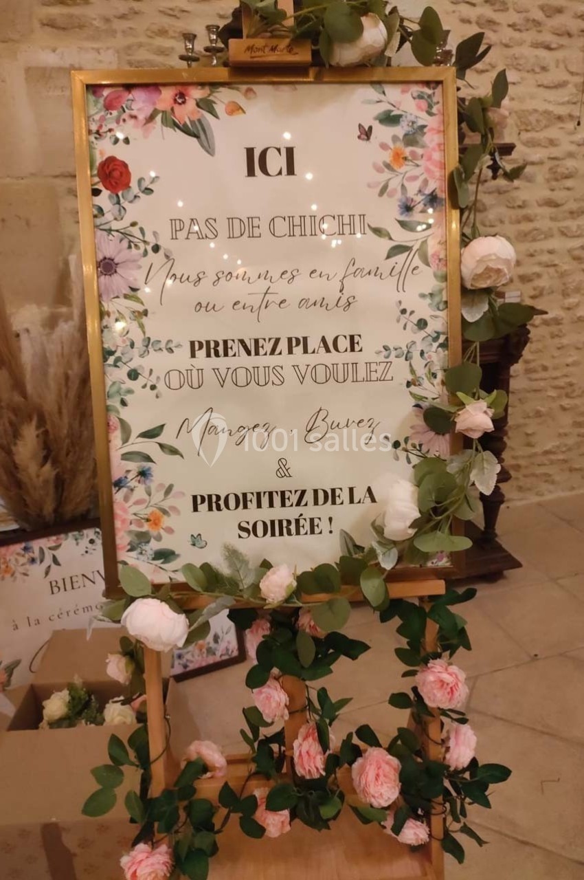 Panneau décoratif avec texte d'accueil entouré de fleurs, posé sur un chevalet dans un décor intérieur en pierre.