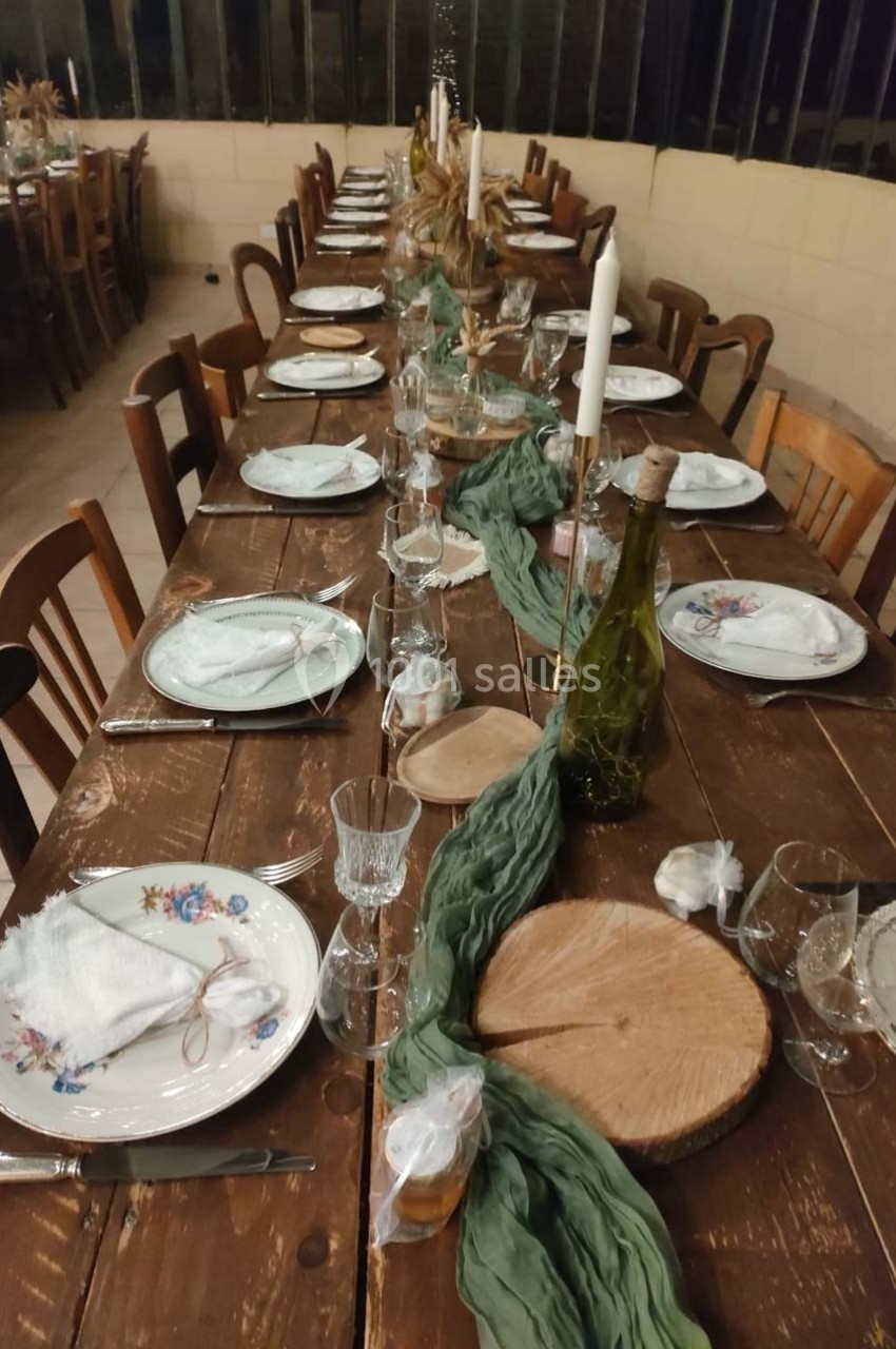 Table en bois rustique dressée pour un repas, avec vaisselle fleurie, bougies, rondins de bois et chemin de table vert.