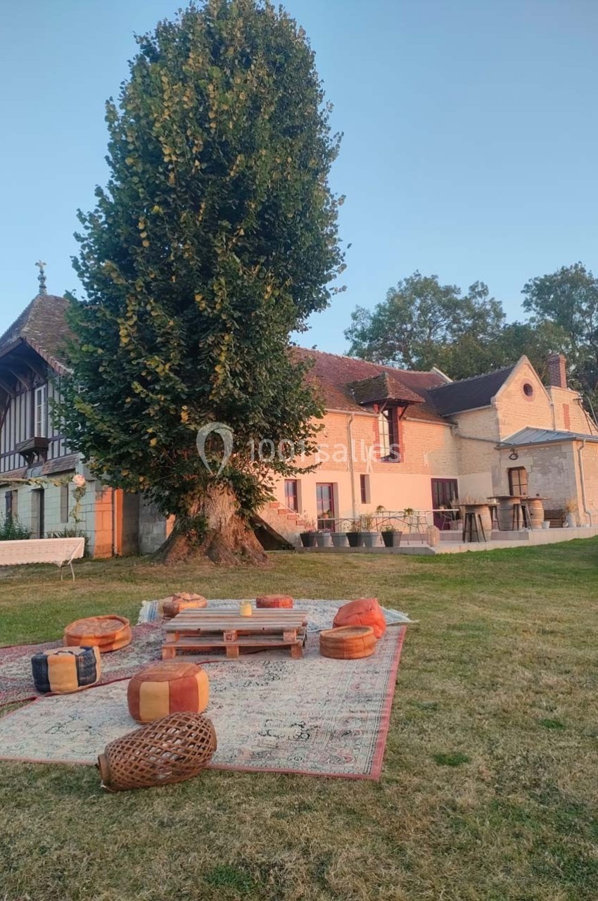 Espace extérieur avec tapis, coussins et table basse en bois devant une grande maison ancienne entourée de verdure.