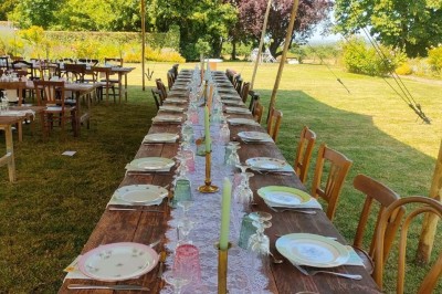 Table en bois rustique dressée pour un repas, avec vaisselle fleurie, bougies, rondins de bois et chemin de table vert.
