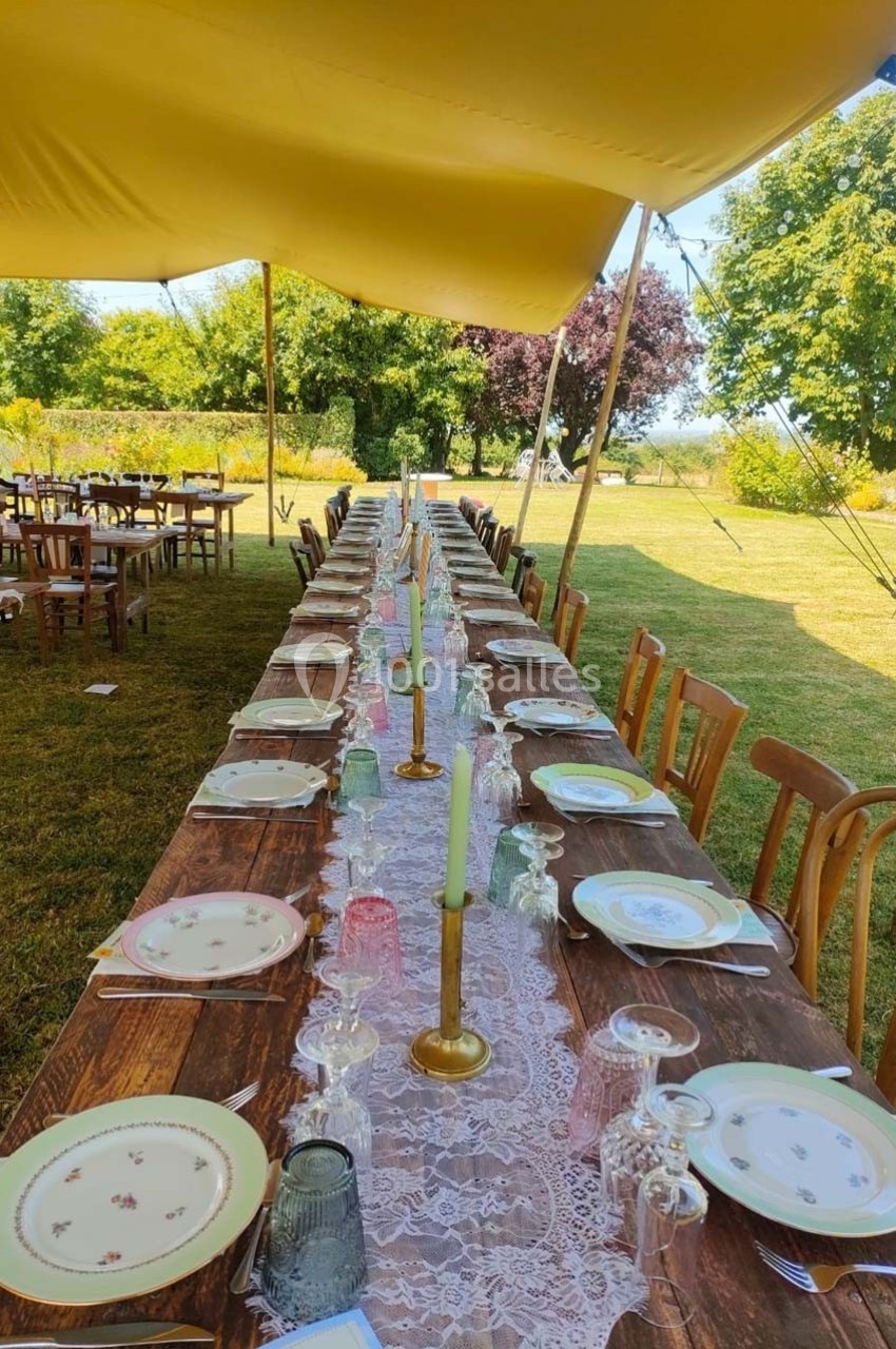 Grande table en bois dressée en extérieur sous une tente, ornée de nappes en dentelle, bougeoirs et vaisselle vintage.