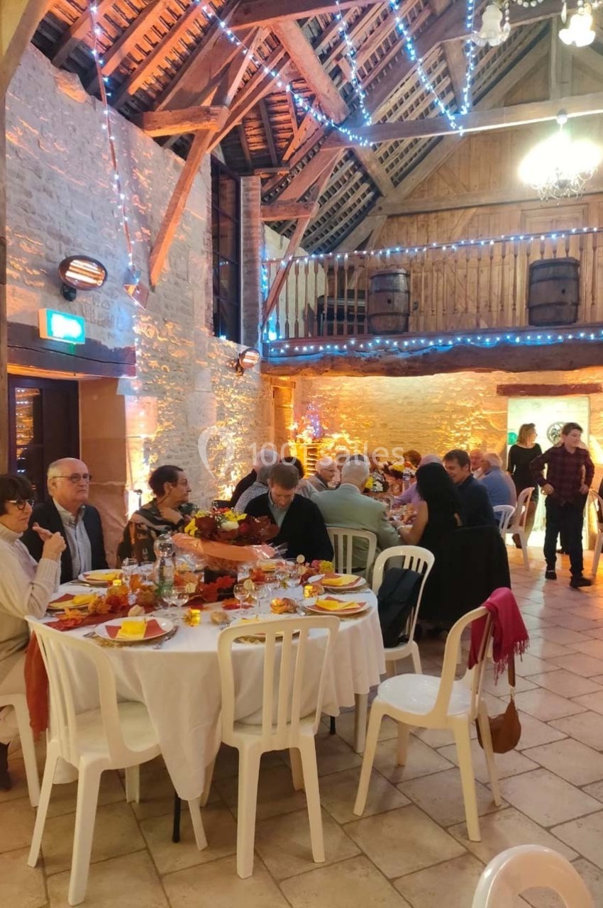 Salle rustique décorée avec des guirlandes lumineuses, des convives assis autour de tables dressées pour un repas convivial.