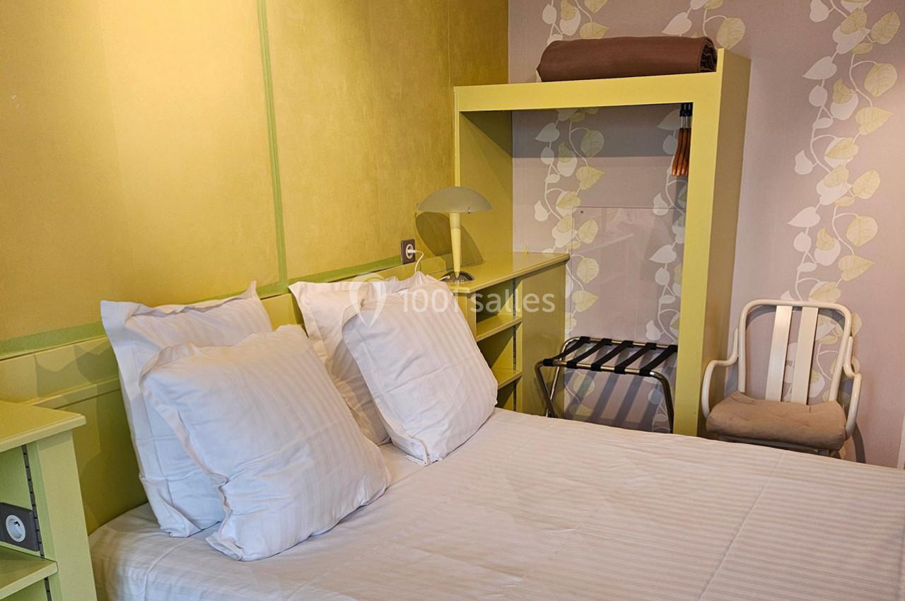 Lit double avec oreillers blancs dans une chambre aux tons jaunes et verts, avec une chaise et un portant en arrière-plan.