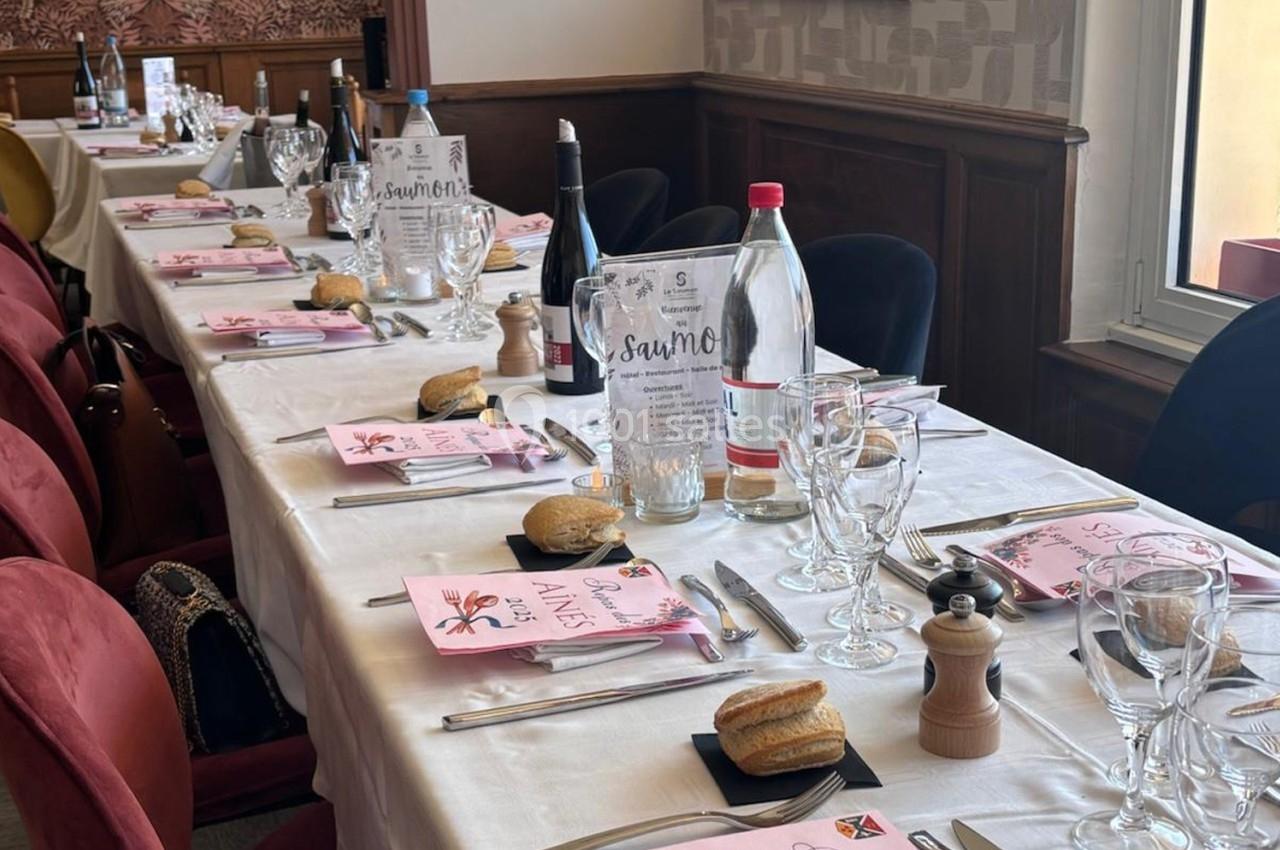 Table dressée pour un repas, avec nappes blanches, assiettes, verres, bouteilles d'eau et de vin, et menus disposés.