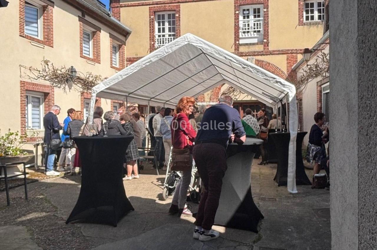 Groupe de personnes discutant sous une tente blanche dans une cour ensoleillée avec des tables hautes noires.