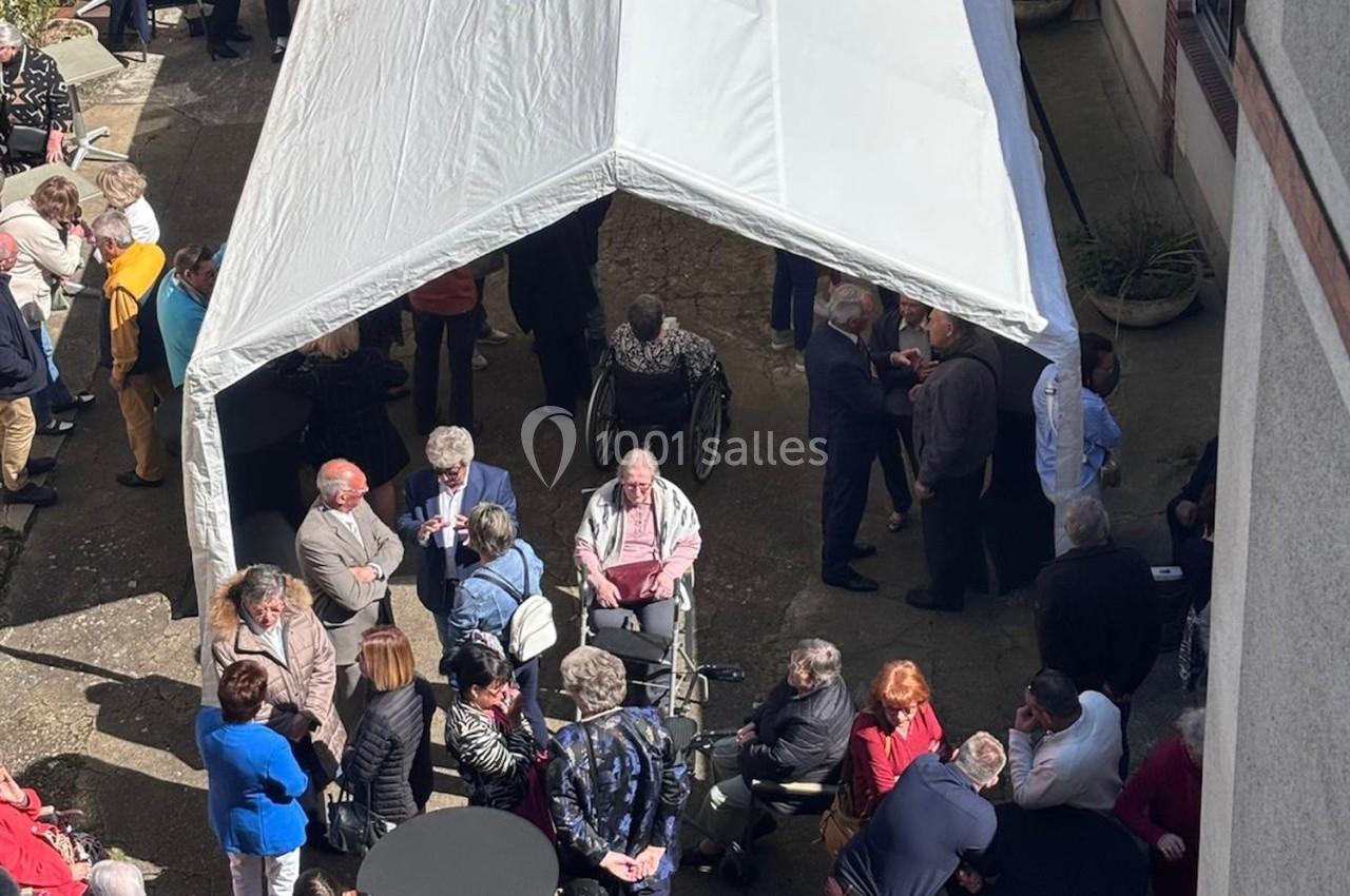 Groupe de personnes rassemblées sous et autour d'un chapiteau blanc dans une cour extérieure, certaines discutant debout.