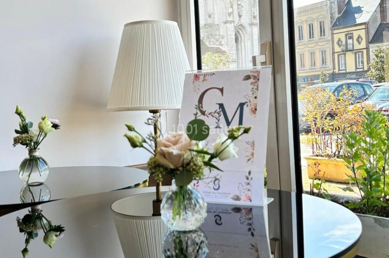 Vase avec des fleurs sur une table en verre, devant une lampe et une affiche, près d'une fenêtre donnant sur la rue.