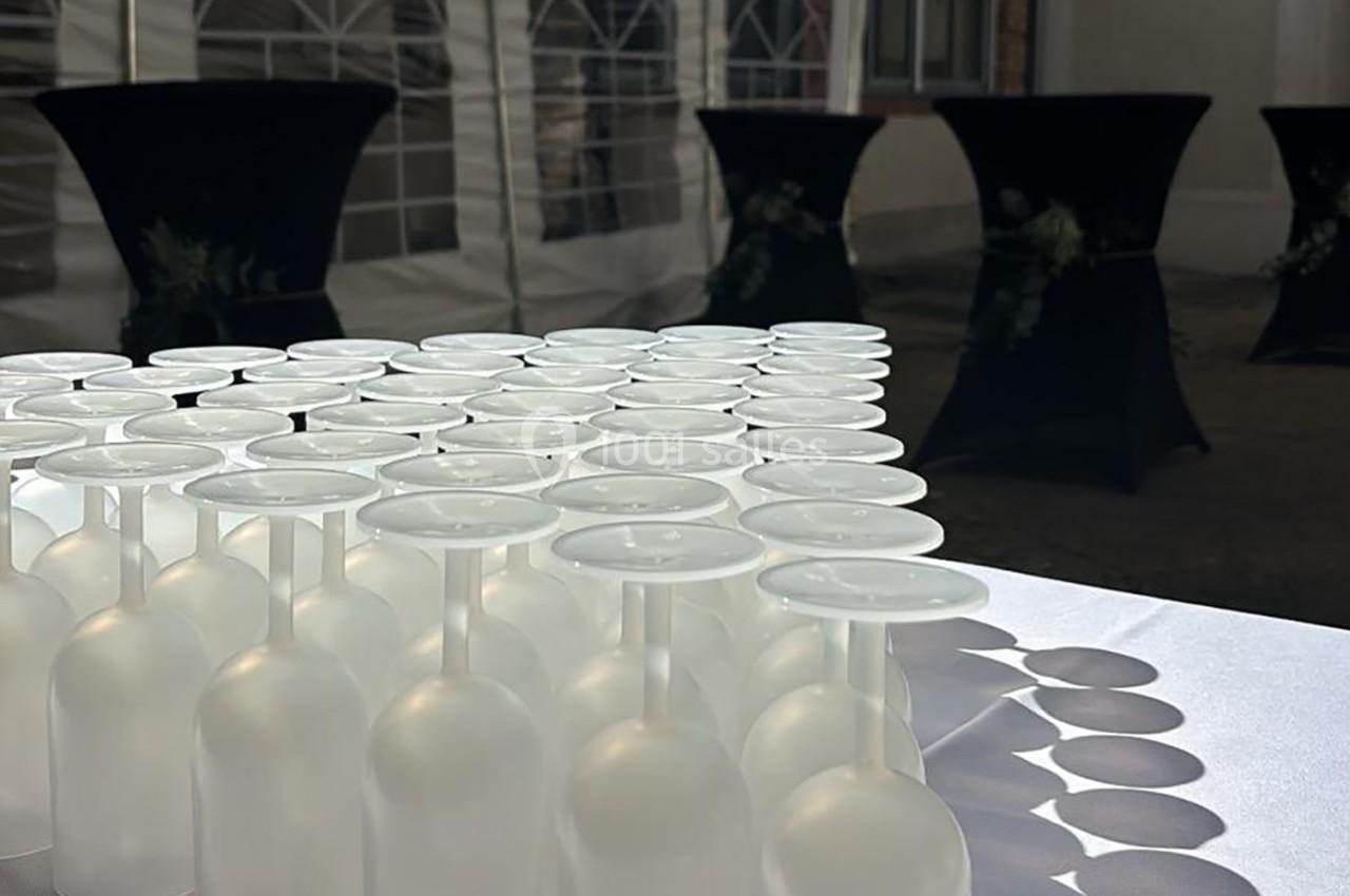 Rangée de verres retournés alignés sur une table blanche, avec des tables hautes en arrière-plan.