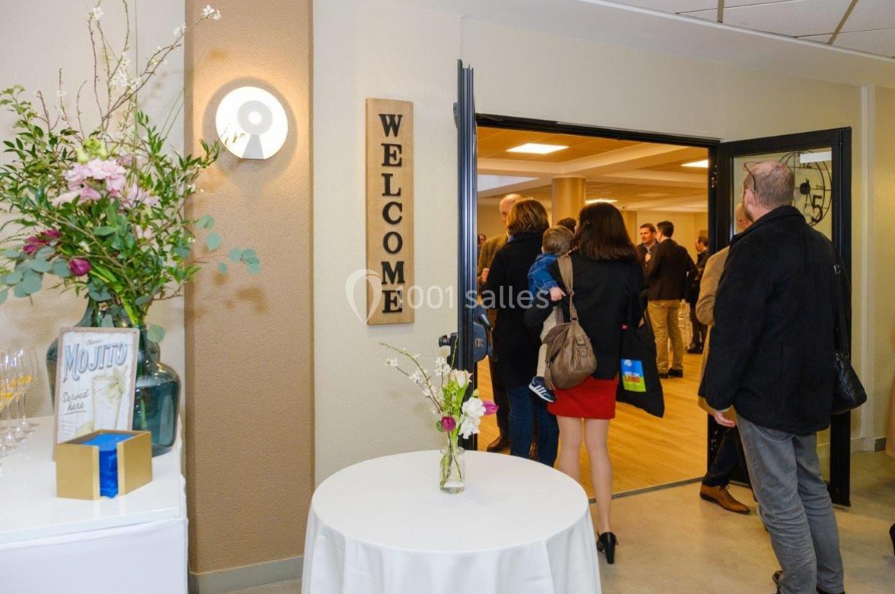 Des personnes entrent dans une salle lumineuse, décorée avec des fleurs et un panneau ’Welcome’ près de l'entrée.
