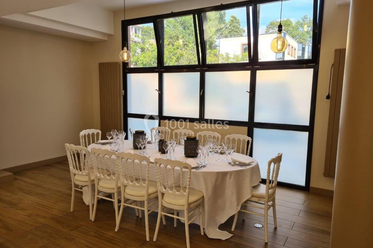 Table dressée avec nappes blanches, chaises élégantes et vaisselle, dans une salle lumineuse avec grandes fenêtres dépolies.