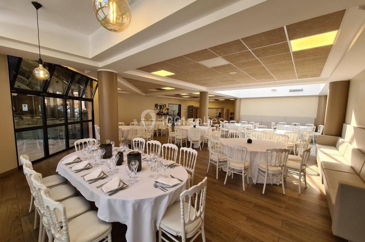 Salle de réception lumineuse avec tables rondes dressées, chaises blanches et décor sobre.