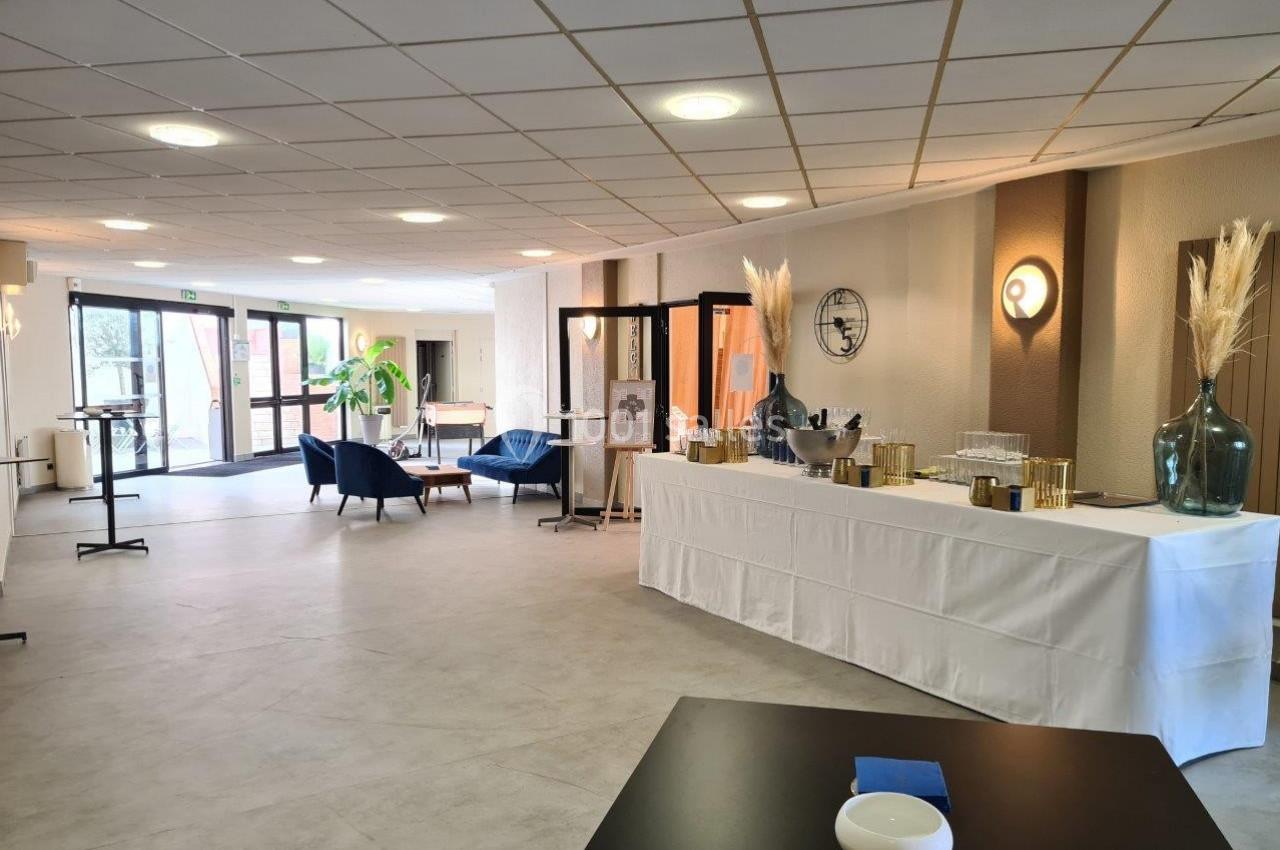 Salle lumineuse avec table dressée pour un buffet, fauteuils bleus et décoration végétale près d'une baie vitrée.
