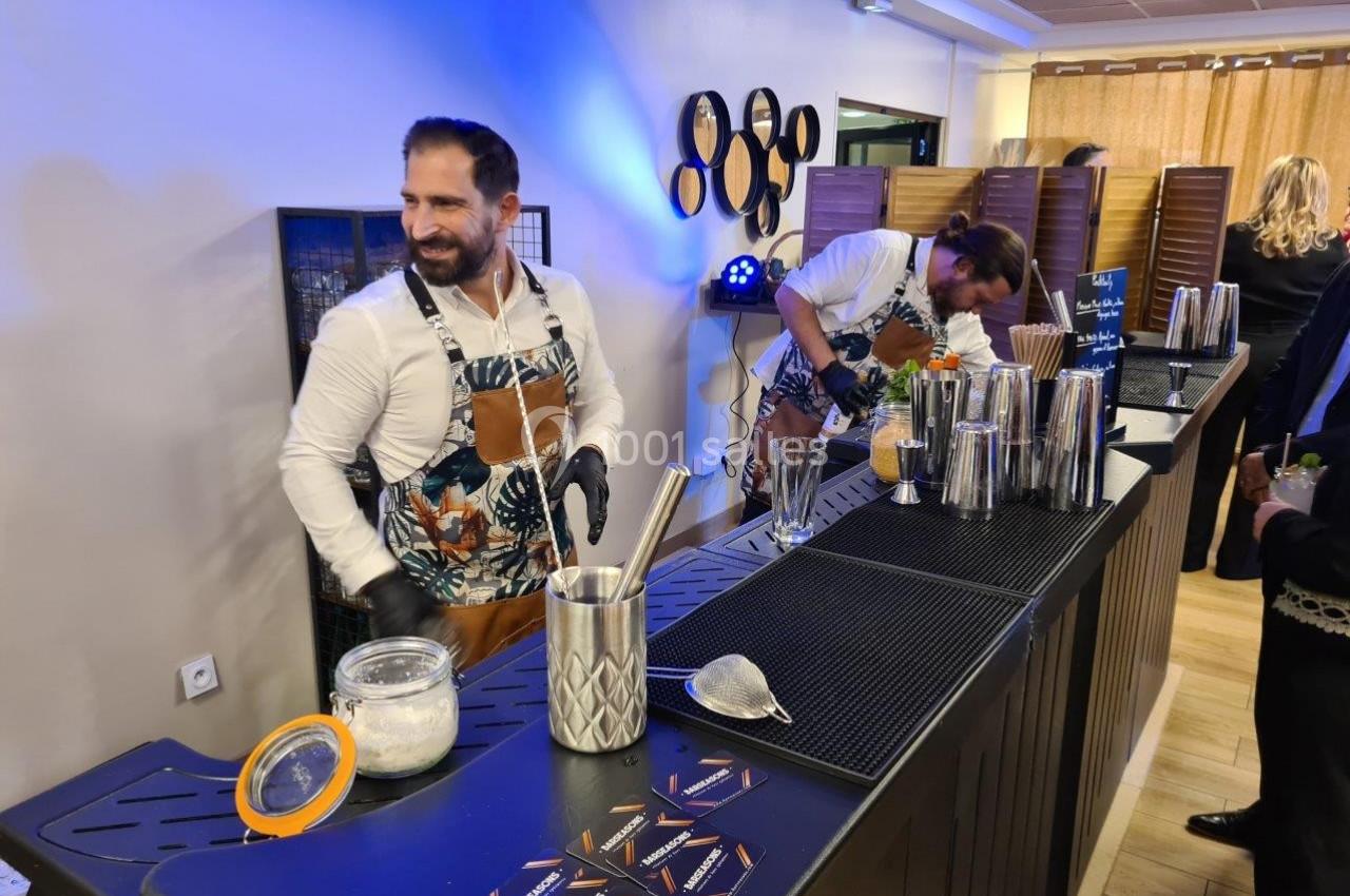 Deux barmans préparent des cocktails derrière un comptoir équipé d'ustensiles et de shakers dans un espace intérieur.