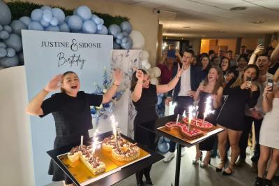 Salle décorée avec des ballons roses, éclairage violet, deux personnes marchant au centre, tables hautes et chaises…
