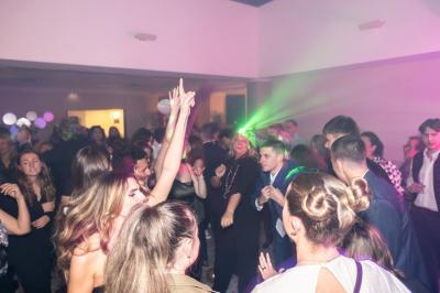 Salle décorée avec des ballons roses, éclairage violet, deux personnes marchant au centre, tables hautes et chaises…