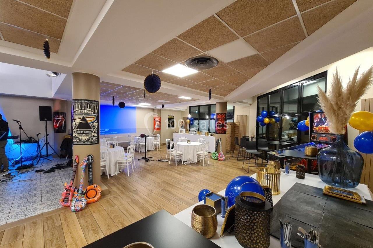 Salle décorée pour un événement avec tables rondes, ballons, affiches rétro et espace pour un groupe musical.