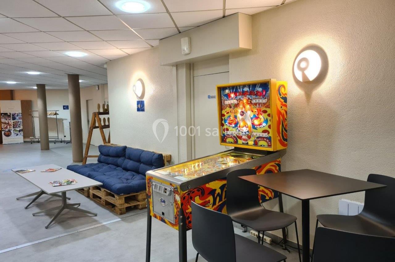 Flipper coloré dans un espace détente avec canapé en palettes, table basse et chaises hautes, éclairage mural.