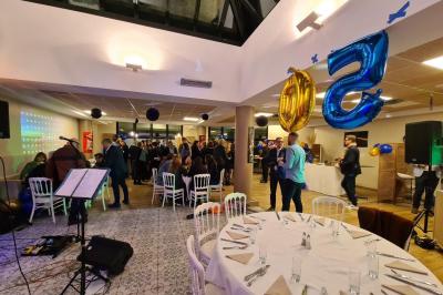 Salle décorée avec des ballons roses, éclairage violet, deux personnes marchant au centre, tables hautes et chaises…