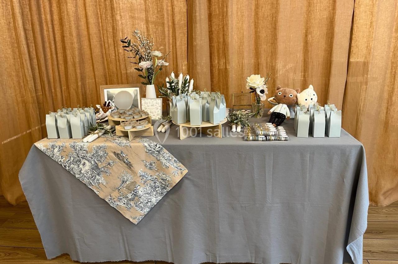 Table décorée avec des cadeaux emballés, fleurs, peluches et nappes, devant un rideau beige.