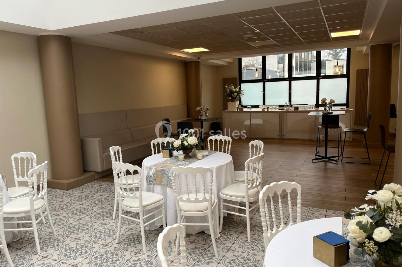 Salle lumineuse avec tables rondes décorées de fleurs blanches, chaises blanches et comptoir en arrière-plan.