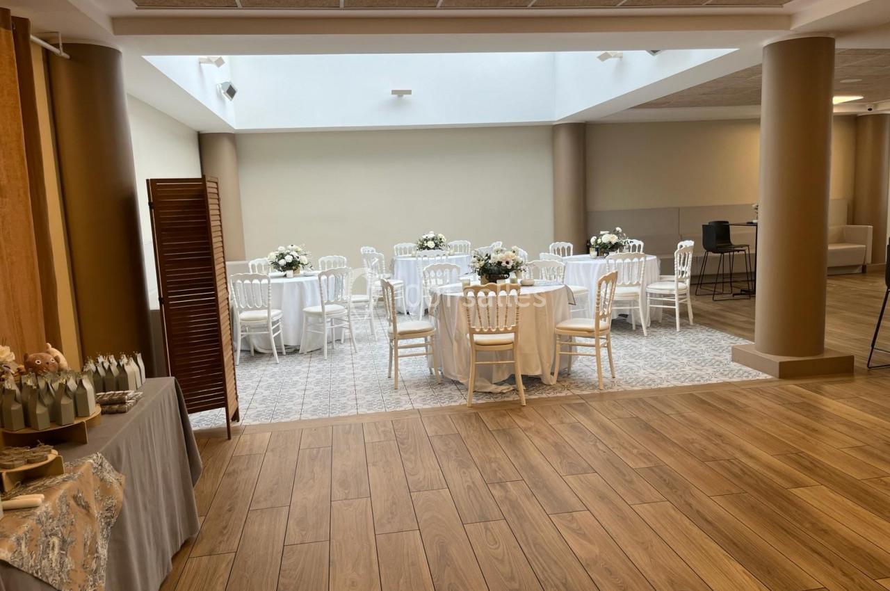 Salle lumineuse avec tables rondes décorées de fleurs blanches, chaises blanches et sol en parquet et carrelage.