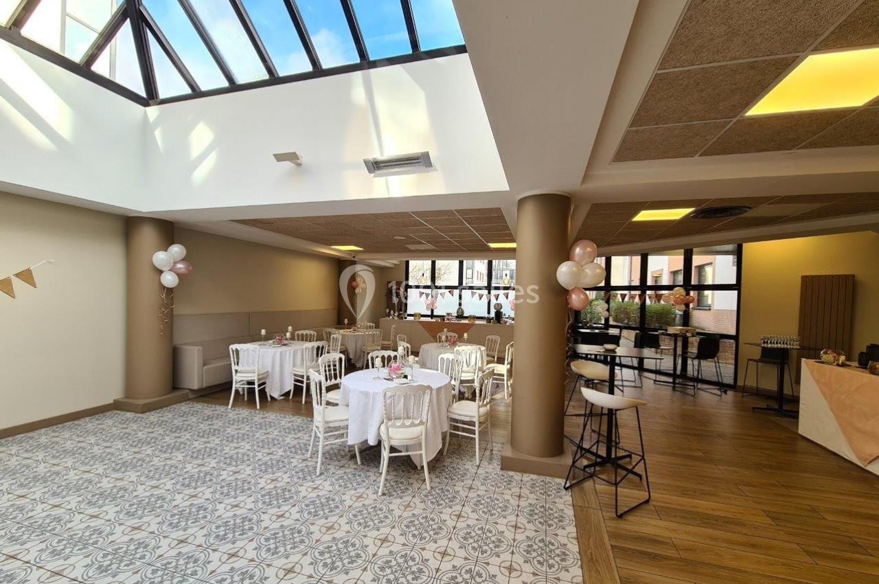 Salle lumineuse avec verrière, tables dressées avec nappes blanches, chaises blanches et décorations festives.