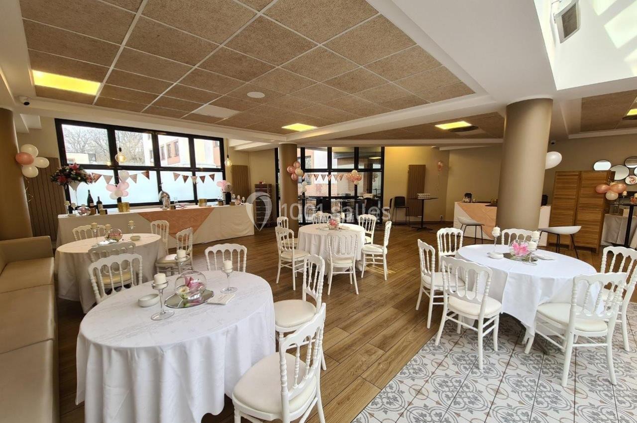 Salle décorée pour un événement, avec tables rondes dressées, chaises blanches et guirlandes suspendues.