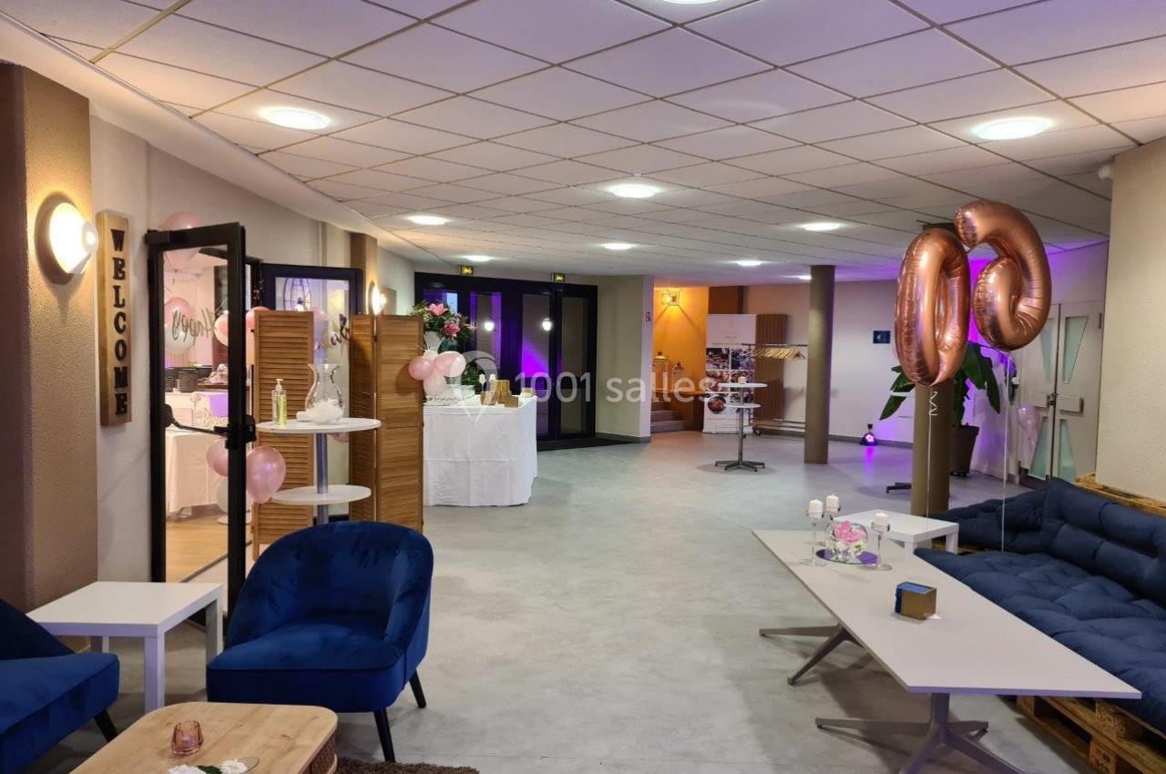 Salle décorée pour un événement avec ballons roses, mobilier moderne et tables ornées de fleurs et accessoires.